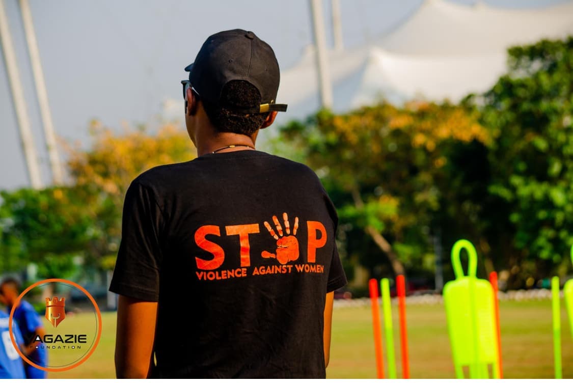 A coach seen from behind in a cap and a black shirt reading Stop Violence Against Women, with training poles in the background