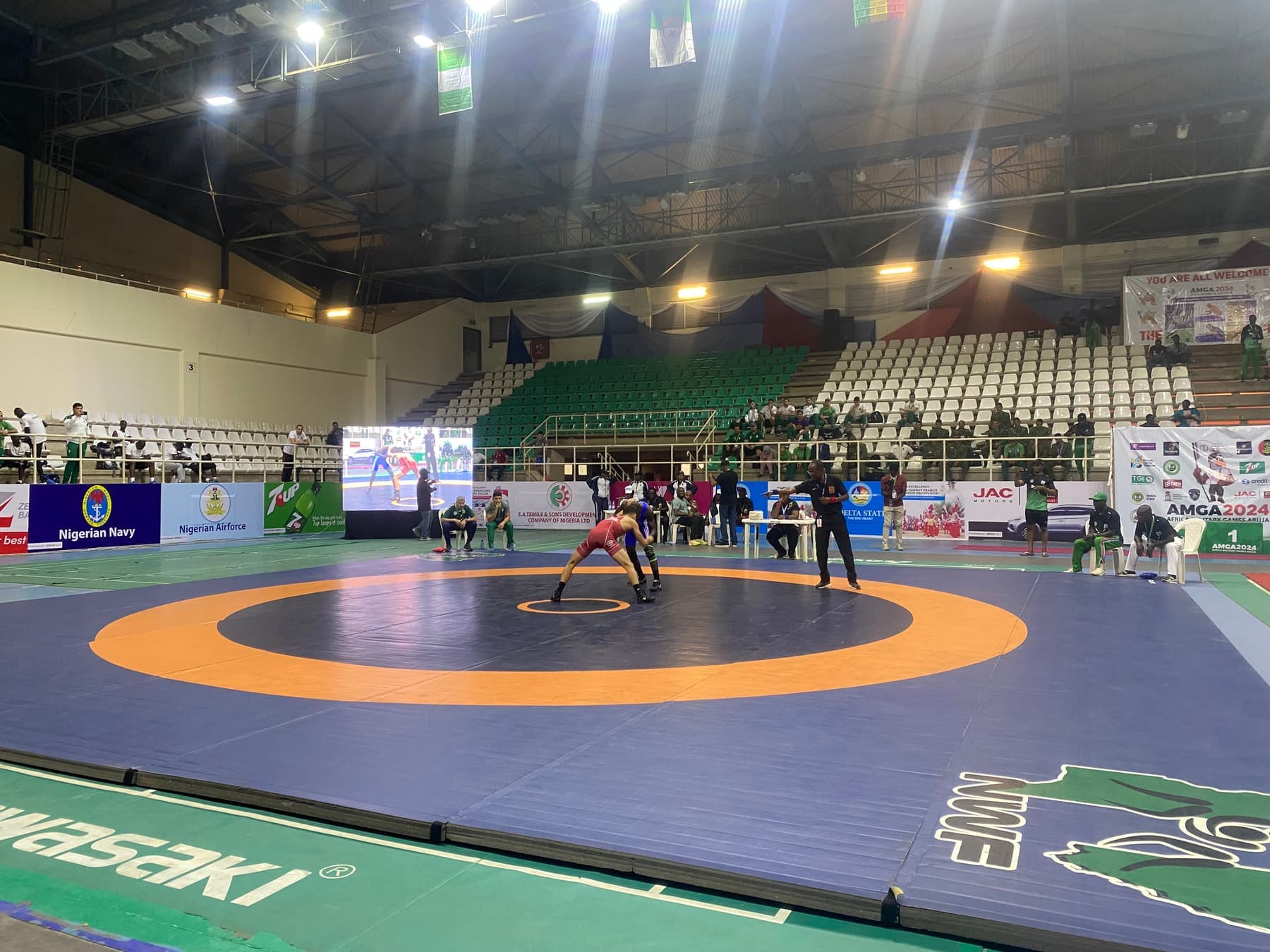 Two wrestlers competing on a mat in an indoor arena watched by officials and spectators