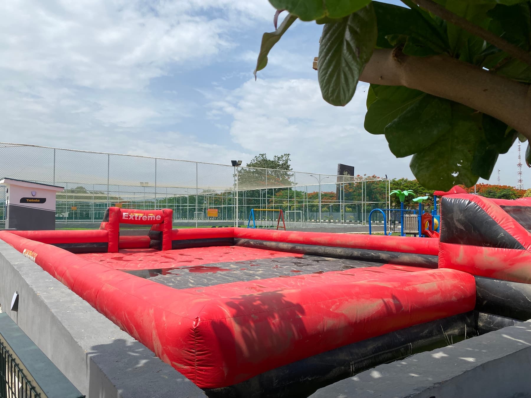 An inflatable red and black arena beside a fenced pitch under a tree