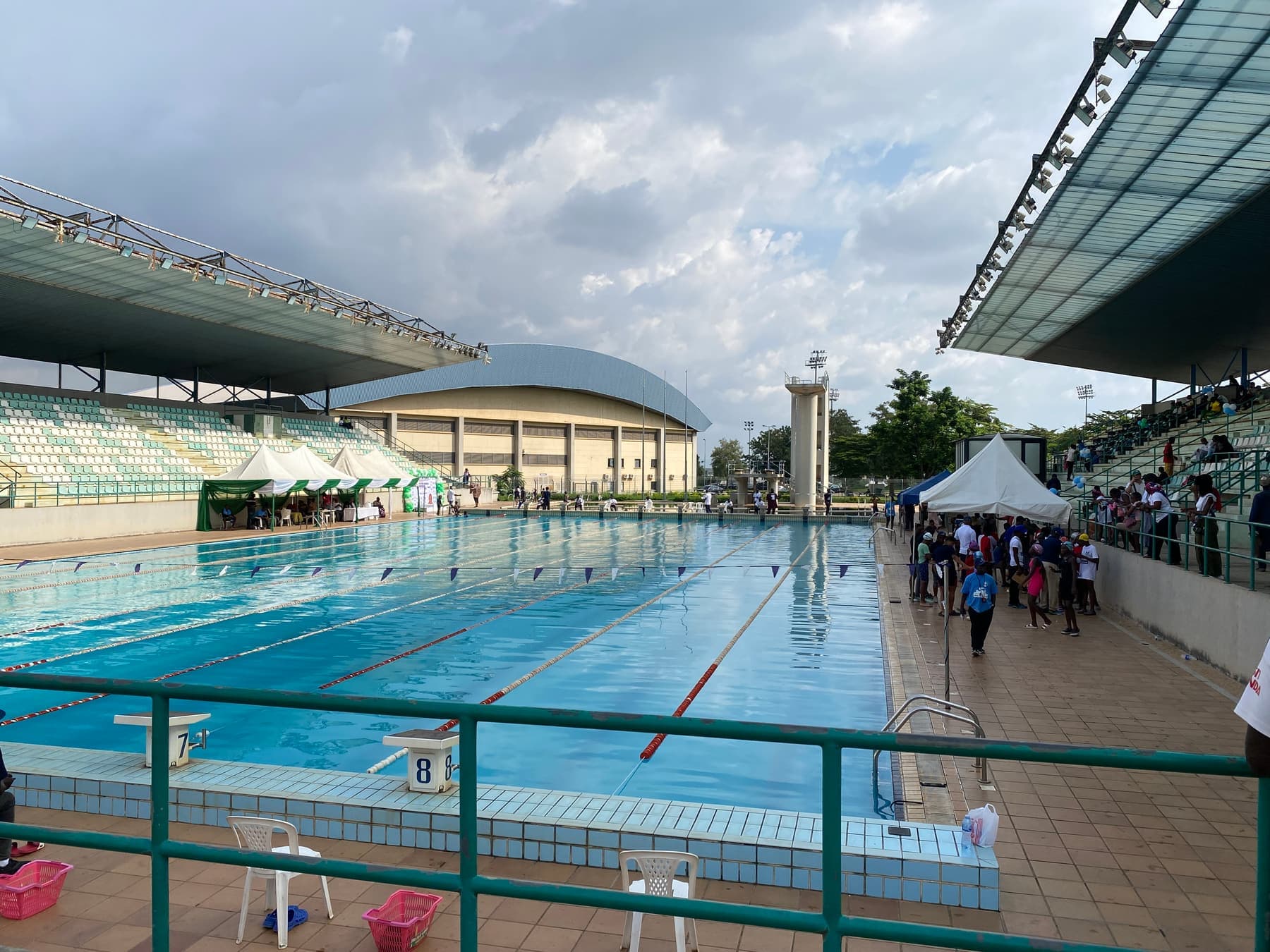An outdoor swimming venue with lane ropes, covered stands and spectators